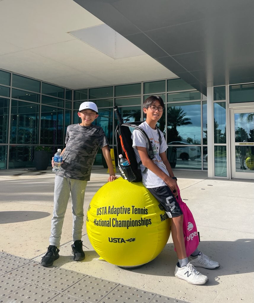 Gavin and Konhee in front of the USTA National campus building. There is a giant tennis ball that says USTA Adaptive Tennis National Championships and Gavin is leaning on it and Konhee is sitting on the giant ball. 